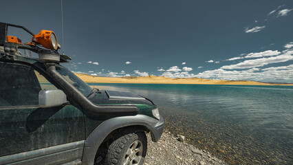 A family off-road vehicle with all-terrain tires parked on the shore of a beautiful lake in the Mongolian desert. Front side view with sand dunes under cloudy blue sky.