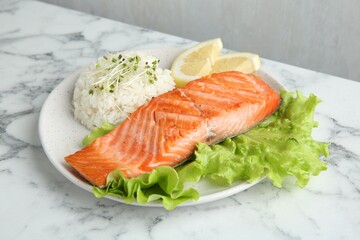 Piece of tasty grilled salmon, lemon, lettuce, rice and microgreens on light marble table against grey background, closeup