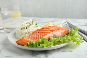 Piece of tasty grilled salmon, lemon, lettuce, rice and microgreens on light marble table against grey background, closeup