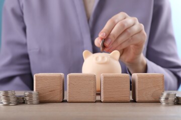 Empty cubes and stacked coins on table, selective focus. Woman putting coin into piggy bank, closeup