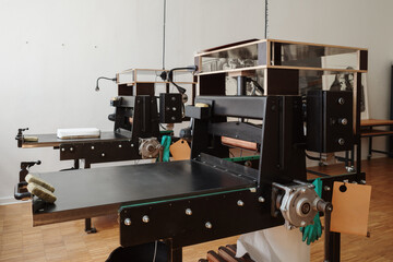 Side view of two professional black etching presses stationed in a bright printmaking workshop with wooden flooring and art supplies.