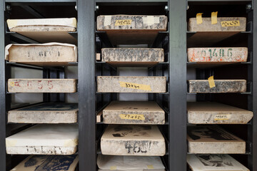 Rows of heavy limestone blocks used for lithography printing stored on black metal shelves. The stones are marked with masking tape and handwritten numbers to organize the inventory in a printmaking.