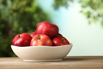 Ripe red apples in bowl on wooden table against blurred green background, closeup. Space for text