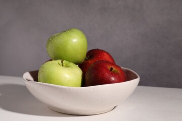 Fresh red and green apples with water drops in bowl on white table against grey background, closeup