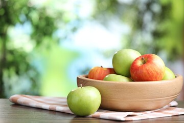 Fresh apples and bowl on wooden table against blurred green background, closeup. Space for text
