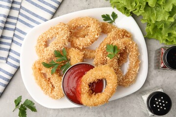Deep fried squid rings with parsley, spices, lettuce and ketchup on grey textured table, flat lay