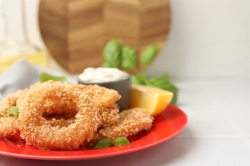 Deep fried squid rings with basil, sauce and slice of lemon on white tiled table, closeup. Space for text