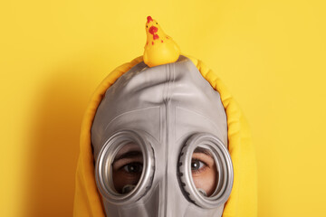 Close-up of a person in a gray gas mask and yellow hoodie with a rubber chicken on top, set against a matching yellow background.