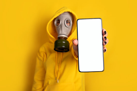 Individual in yellow hoodie and gas mask holding smartphone with blank screen, isolated on vibrant yellow background, concept of technology and protection.