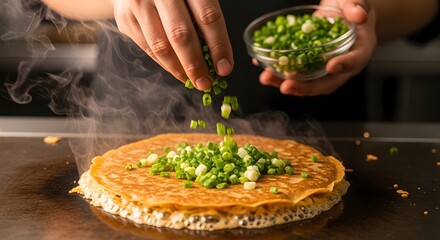 Chef's hands sprinkling fresh chopped green onions onto a sizzling savory pancake on a hot griddle, creating a delicious meal.