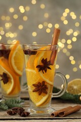 Delicious mulled wine with slices of orange, spices and fir branches on wooden table against grey background with blurred Christmas lights, closeup. Bokeh effect