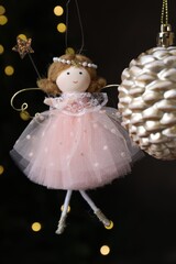 Fototapeta premium Beautiful Christmas bauble and fairy doll hanging against black background with blurred lights, closeup
