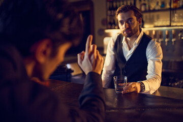 Customer gesturing toward bartender holding glass in warm bar lighting. Concept of ordering interaction, beverage choice moments, bar communication, and drink-focused service atmosphere.