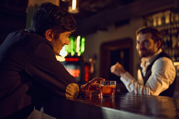 Man chatting with bartender while resting cocktail on bar counter. Concept of social drinking, relaxed service moments, beverage enjoyment, and warm bar ambiance.