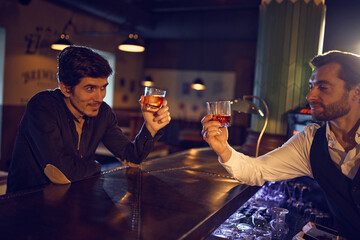 Bartender and guest clinking whiskey glasses at bar counter. Concept of bar hospitality visuals, drink service marketing, social ritual moments, and alcohol brand promotion content.