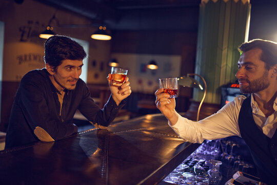 Bartender and guest clinking whiskey glasses at bar counter. Concept of bar hospitality visuals, drink service marketing, social ritual moments, and alcohol brand promotion content.