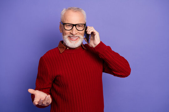 Confident senior man with red sweater on purple background talking on smartphone smiling and inviting conversation for advertising and lifestyle use