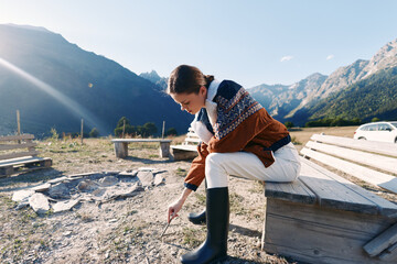 Fototapeta premium Woman in sweater and boots sits on a bench by a campsite in the mountains, playing with a stick near a fire pit, sunlight and distant peaks create a peaceful outdoor nature travel scene.