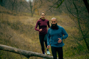 Couple running past fallen log during relaxed forest training. Concept of easy trail cardio, daily habit, outdoor fitness, calm motion, natural wellness routine.