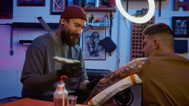 Medium shot of focused young bearded Caucasian male tattooist in stylish beanie hat and sterile gloves using tattoo machine while drawing body art on male clients arm in professional studio