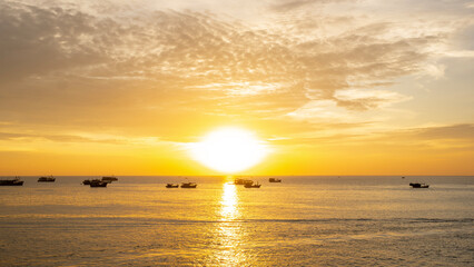 A warm golden sunset over the sea, with several small boats silhouetted against the glowing horizon and soft clouds scattered across the sky