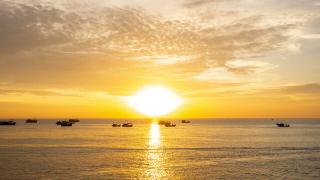 A warm golden sunset over the sea, with several small boats silhouetted against the glowing horizon and soft clouds scattered across the sky