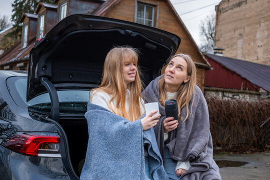 Mother and daughter wrapped in cozy blankets, enjoying warm drinks together by the car, sharing joyful moments and strengthening their family bond in a serene outdoor setting - Powered by Adobe