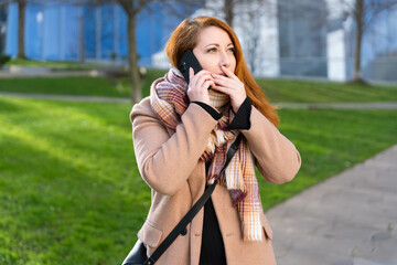Redhead woman having a surprising phone call, expressing shock and concern in an urban park during autumn