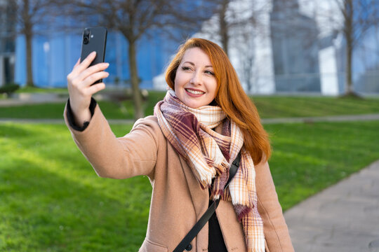 Redhead woman taking selfie with smartphone in urban park, smiling for social media content outdoors