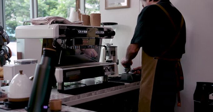 Asian senior male barista preparing fresh espresso using professional coffee machine inside small cozy cafe during retirement with focus on portafilter placement and manual brewing motion
