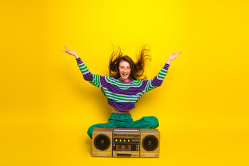 Energetic young woman with striped sweater sits on a vintage boombox against a bright yellow background expressing joy and fun in a casual fashion shoot