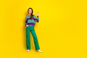 Young female model in striped sweater and green pants waves against a bright yellow background