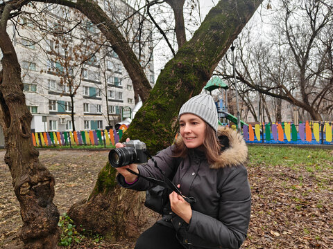 Young woman with a camera, sitting on the ground beside a tree, capturing images in a colorful park, showcasing creativity and passion for photography in a vibrant environment - Powered by Adobe