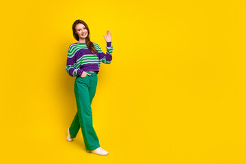 Young female model wearing colorful striped sweater green pants smiles waves yellow background for a cheerful fashion lifestyle promotional image