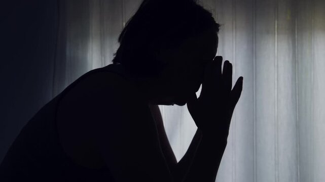 Silhouette of sad woman sitting by window with head in hands, feeling depressed and lonely at home. 4K.
