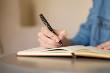 Close up of a woman's hand writing in notebook with a pen. The young female student is studying, working, journaling, taking notes, writing while sitting at desk with a blurred background. 