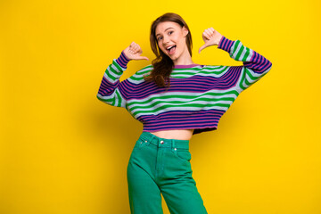 Young woman posing in colorful striped sweater and green pants against yellow background smiling...