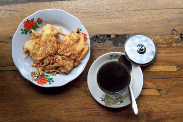 Top-down view of fried banana fritters on a vintage floral enamel plate next to a glass of black coffee on a rustic wooden table. Traditional Indonesian snack setting.
