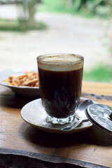 Glass of traditional black coffee on a rustic wooden table with a metal lid and spoon. Blurred background features a plate of fried snacks and a green garden view. Authentic rural Indonesian setting.