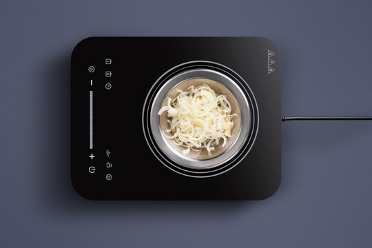 On a sleek, modern induction cooktop, thinly sliced onions are cooking in a metallic pot. The kitchen has a contemporary design with a minimalist vibe, focusing on simplicity.