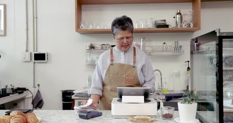 Asian senior woman working behind point of sale system in cozy coffee shop with croissants and cup on counter showing dedication to small business operation during retirement lifestyle
