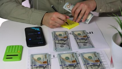 Close-up of an investors hands adding income annotation stickers to organized money stacks alongside IPO research materials. This detailed financial organization scene showcases meticulous investment