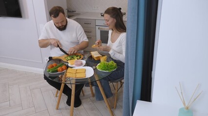 Happy couple making sandwiches for breakfast or lunch in their modern kitchen, slicing cucumbers and spreading butter on bread while enjoying time together