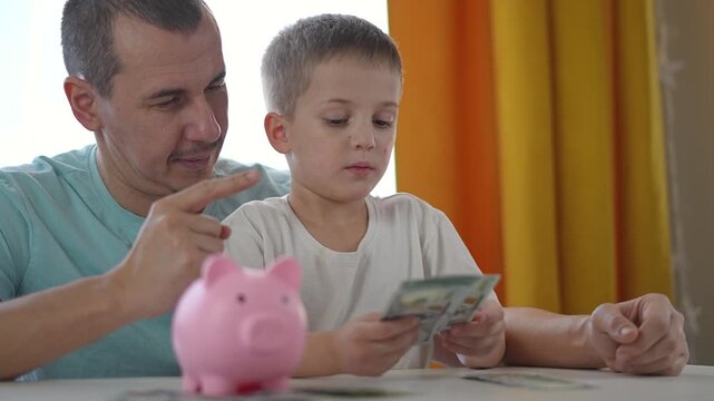 Boy counts dollar bills while father sits beside him and explains saving basics. Repeated money handling builds child finance skills. Saving taught step by step in natural home environment.