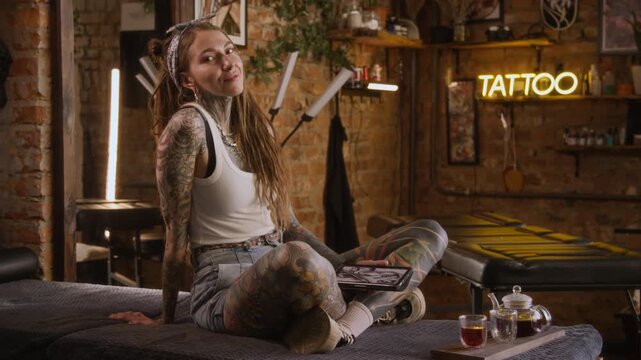 Full portrait of confident young Caucasian female tattoo artist with full body tattoo posing for camera in modern tattoo parlor with yellow neon sign while working on digital sketch on tablet