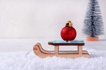 A red Christmas tree ball on a toy wooden sled in artificial snow. A spruce tree in the background...