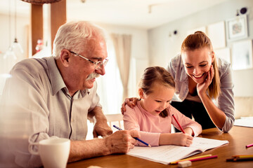 Senior grandfather, adult mother, and young daughter happily doing homework at home