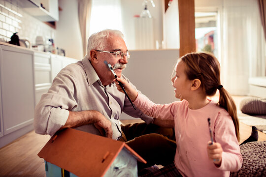 Senior grandfather and child granddaughter laughing while painting at home