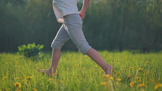 Running child in motion across grassy summer field. Foot leaves ground in barefoot stride. Nature around moves as child runs through motion of summer light. Legs of child show barefoot summer energy.