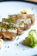 Close-up of pork jelly slices garnished with chopped chives served on a plate with cucumber garnish in a bright setting